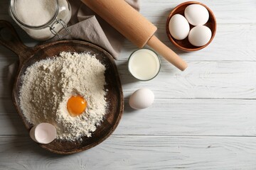 Pile of flour with yolk and other ingredients for dough on white wooden table, flat lay. Space for text