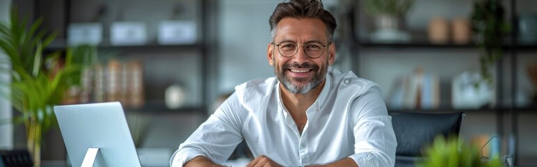 Obraz premium A happy businessman sitting at his desk in an office, smiling and looking into the camera with glasses on. He is wearing white shirt Smiling Businessman in Modern Office with Laptop - 4K HD Wallpaper