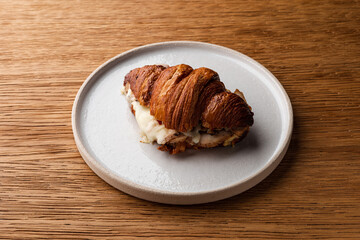 Delicious Breakfast Croissant Sandwich on a White Plate: Flaky Golden-Brown Croissant Filled with Scrambled Eggs, Ham, Cheese, and Fresh Vegetables, Served on a Wooden Table - Side View