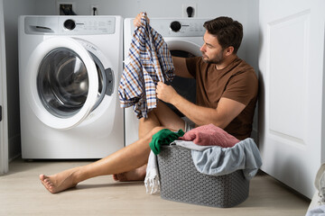 Happy man sorting clothes near washing machine in laundry room. man sits on the floor of a house...