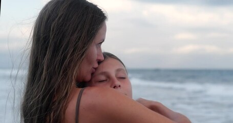 Portrait of a mother and her teenage daughter standing on the beach near the ocean during sunset. A woman kisses her daughter on the forehead and hugs her while looking out at the ocean. Mothers Day.