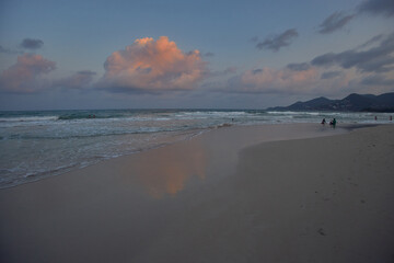 Beach scene at dusk with a vibrant cloud reflection on wet sand and distant figures by the water