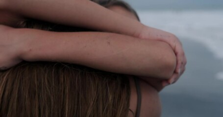 Close-up shot of a teenage girl hugging her mom tenderly and lovingly on the beach near the ocean. She hangs on her mother's neck and looks up at the sky. Mothers Day. The affection of a child.