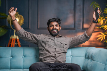 Excited Indian man sitting on sofa and rising his hand with mobile phone