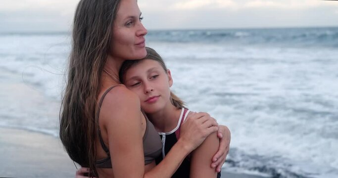 Portrait of a mother and her teenage daughter standing on the beach near the ocean during sunset. A woman kisses her daughter on the forehead and hugs her while looking out at the ocean. Mothers Day.