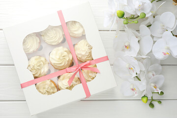 Tasty cupcakes with vanilla cream in box and orchid flowers on white wooden table, top view