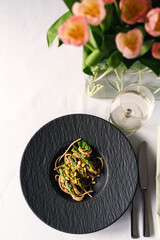 Spaghetti mit grünem Spargel, Pinienkernen und Zitrone in schwazen Tellern auf Tisch mit weißer Tischdecke und Tulpen Blumenstraß