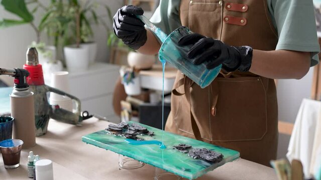 Female craftswoman working with epoxy resin in liquid art technique. Hands of woman artist in gloves pouring viscous blue fluid from plastic cup, creating making unusual water pattern on art project.