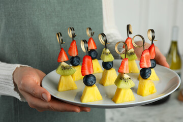 Woman holding plate of tasty canapes with pineapple, kiwi and berries indoors, closeup