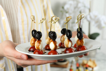 Woman holding plate of tasty canapes with black olives, mozzarella and cherry tomatoes indoors,...