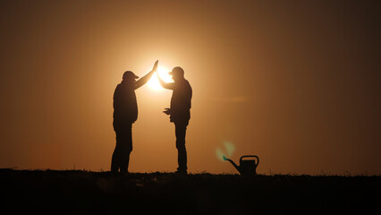 Making a high five gesture, silhouettes of two successful farmers stand in a field.