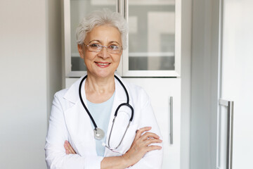 Happy successful senior female doctor in white uniform and stethoscope standing with folded arms in her cabinet at hospital or private clinic, reminding you to take care of your health