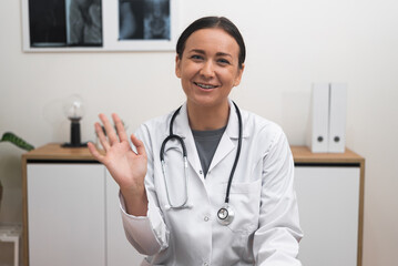 Online Consultation Welcome: With a welcoming smile, a female doctor waving hand engages with the camera, making patients feel comfortable during their online appointment. 