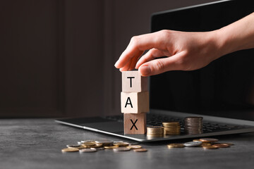 Woman with word Tax made of wooden cubes, laptop and coins at grey table, closeup. Space for text