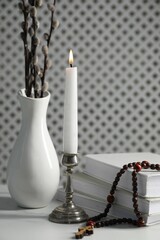Books, burning candle, rosary beads and vase of willow branches on white table, closeup
