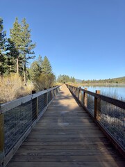 A wooden boardwalk winds along a serene mountain lake, framed by towering peaks. The bridge invites a leisurely stroll, offering breathtaking views of the tranquil waters and rugged landscape.