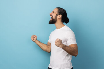 Side view of excited man with beard wearing white T-shirt expressing winning gesture with raised...