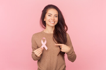 Portrait of adorable smiling woman with wavy hair, holding pointing at pink ribbon, symbol female...