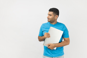 Portrait of handsome unshaven man wearing blue T- shirt standing holding closed laptop, looking...
