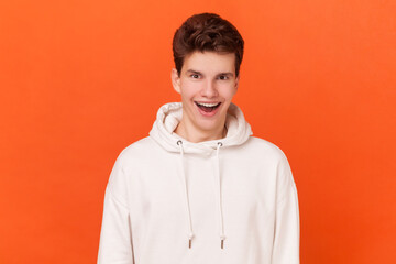 Portrait of astonished young handsome man wearing white hoodie looking at camera with open mouth,...