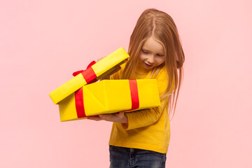 Happy satisfied blonde little girl holding big yellow gift box, looking inside being glad to get...