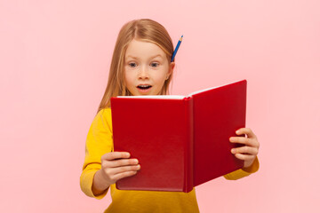Portrait of amazed surprised blonde little girl reading book with excited face, being inspired with fairy tale plot, wearing yellow jumper. Indoor studio shot isolated on pink background.