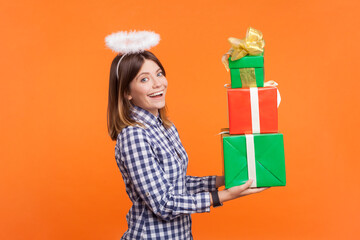 Profile portrait of happy adorable woman with brown hair and nimb over head holding stack of gift boxes, wearing checkered shirt. Indoor studio shot isolated on orange background