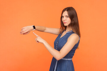Portrait of strict confident serious brunette woman wearing denim dress pointing at her wristwatch...