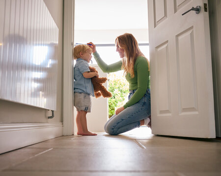 Mother Measuring Height Of Young Son Holding Teddy Bear At Home Against Kitchen Door Frame