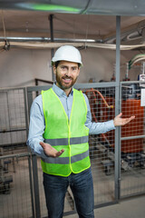 Confident joyful man architect in a hard hat looks at the camera and shows installations inside plant