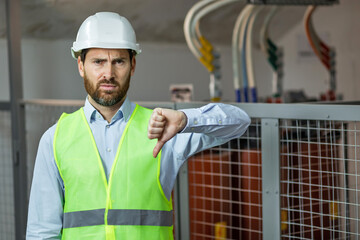 Sad man in a safety vest and helmet gives a thumbs down with negative expression, electrician...