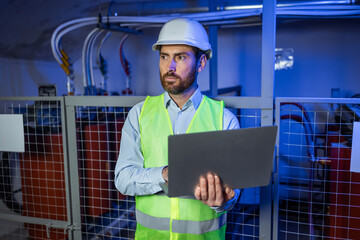 Engineer with laptop in basement of industrial building. Industrialist man in protective helmet. Factory employee.