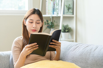 Having relaxing weekend, comfort asian young woman with cup of beverage, drinking coffee or tea of mug. Girl sitting on sofa open reading book enjoy of rest after get up in morning lifestyle at home.