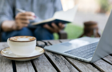 Close-up shot of coffee cup with laptop on old wooden table, blurred a man taking a note on...