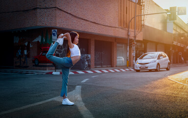 A woman performs a stretching pose in an urban street at sunset, showcasing flexibility and...