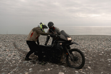 Friends bikers helping in trip on adventure motorcycle towing on sea shore pebbles
