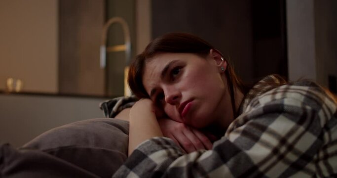 A calm and bored brunette girl in a checkered shirt leans on a pillow on a gray sofa she is bored and looks away in a modern apartment in the evening