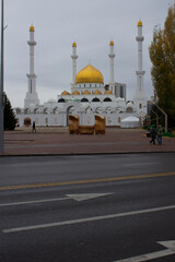 Vertical street photo. The Nur-Astana Mosque with golden dome, four minarets, white wall. Third largest mosque in Central Asia. Asphalt roadway with white road markings, arrow. Kazakhstan. Islam. Day - obrazy, fototapety, plakaty
