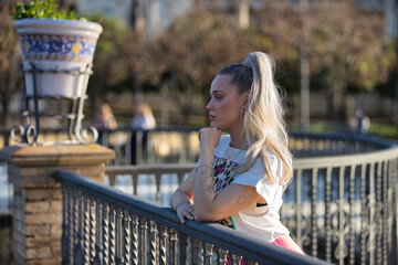 Young, pretty, blonde woman, with blue eyes and a printed T-shirt, leaning against a railing,...