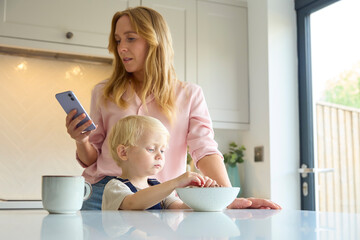 Mother At Home Looking At Mobile Phone As Young Son Eats Breakfast In Kitchen