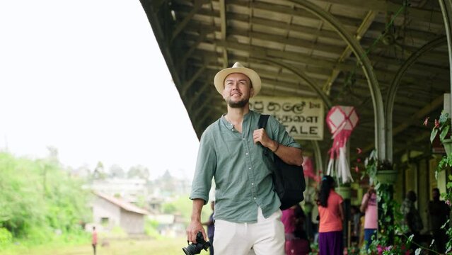 Asian travel blogger explores Haputale railway station with camera, backpack. Man in hat walks platform, records train trip. Authentic Sri Lanka station, voyager captures local culture, adventure.