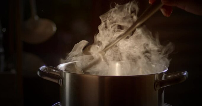 Slow motion scene of a boiling pot steams up as a restaurant's chef uses a ladle to stir soup in a cooking pot in the morning.


