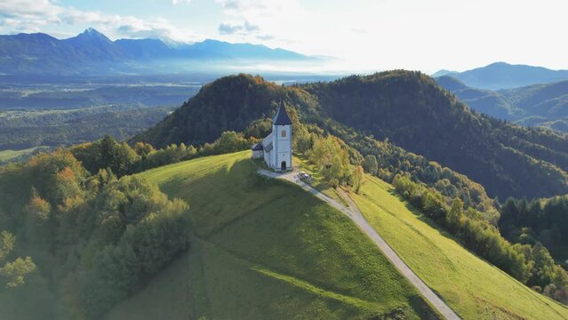 View from above of the serene beauty of the small Church of St. Primoz in Jamnik caressed by the fog at sunrise on green hill in Slovenia. Julian Alps, Kranj, Gorenjska