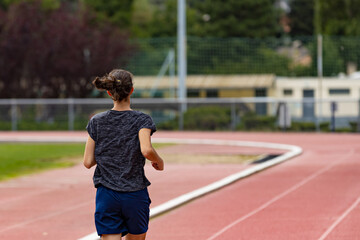 Fototapeta premium Back view of a young woman running on a stadium track; back view of a young woman jogging on a running track; action shot of an athlete warming up .