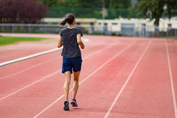 A young woman with a ponytail jogging on a stadium track in shorts and a t-shirt; a female athlete running along a track on a sunny day; back view of a female athlete training on a running track.