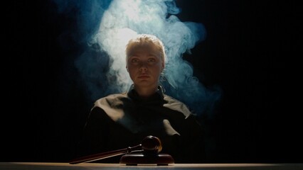 Female sitting at the desk with wooden hammer, dark room bright spotlight and smoke. Woman judge...