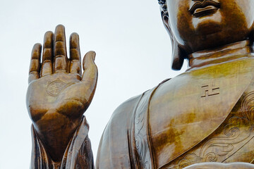 The image is a statue of Buddha, made of bronze, with his right hand raised in the Abhaya mudra, a...