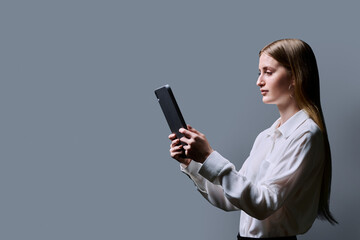 Teenage female student using digital tablet on gray studio background, profile view
