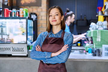Young woman service worker in apron looking at camera in restaurant, coffee shop