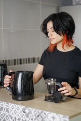 Black-haired white person smiling while making a coffee. They are holding a water heater and there...